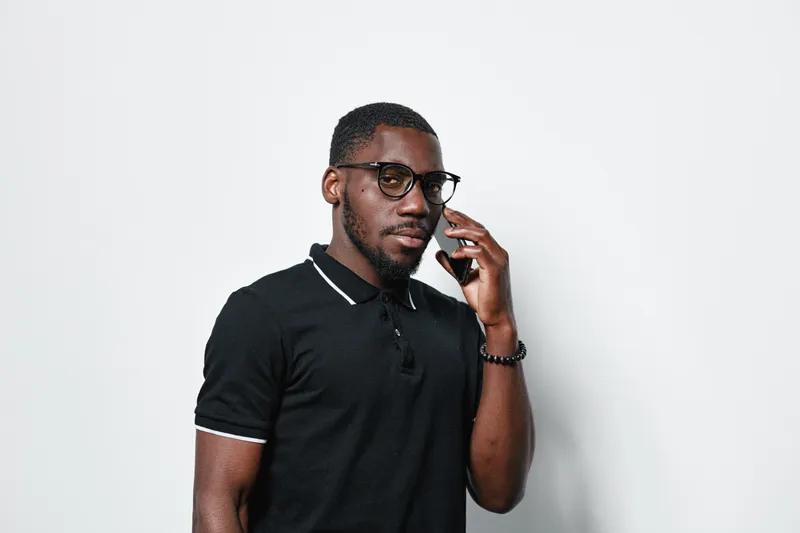 African American man with dark skin and glasses talking on smartphone wearing black polo shirt standing against white background. Serious expression and casual style.