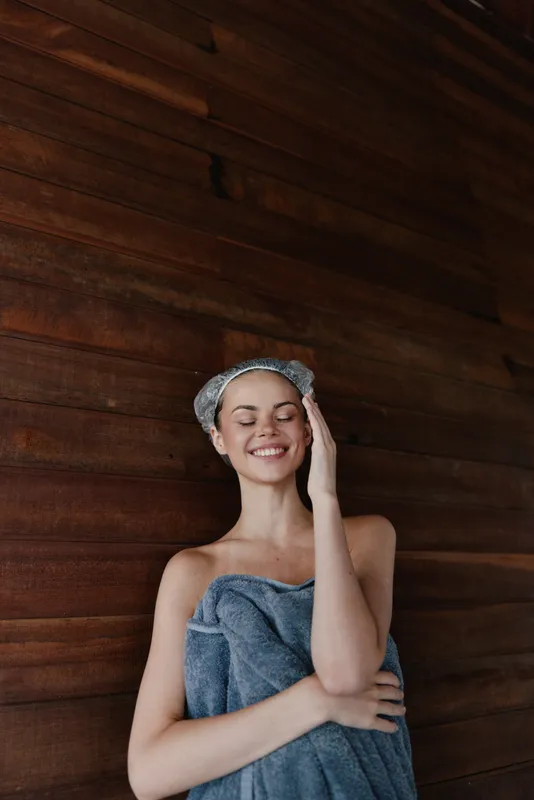 smiling woman wrapped in blue towel enjoys spa moment with eyes closed against wooden wall, feeling relaxed and peaceful indoors after bath