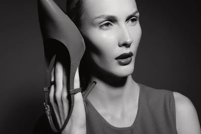 Elegant white woman with pale skin holds high heel shoe near face in black and white studio portrait.