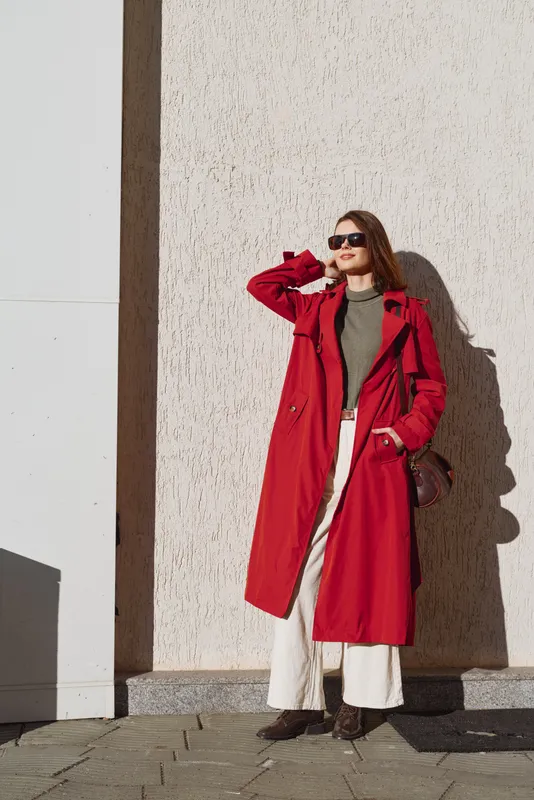 Young woman wearing a long red coat and sunglasses stands confidently against a textured wall on a sunny day, expressing style and urban fashion.