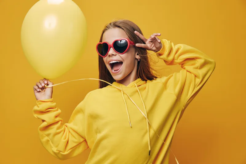 Happy woman in yellow hoodie holding balloon and wearing red heart sunglasses on yellow background, smiling and making peace sign with fingers for celebration and fun.