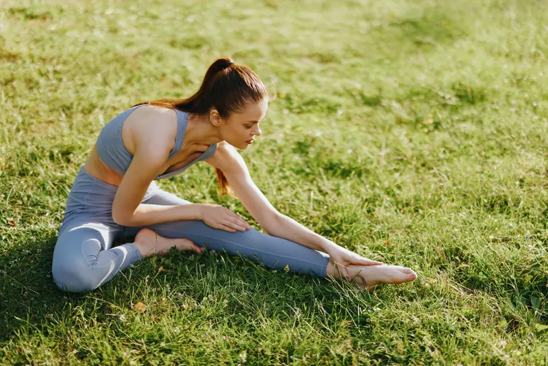 Young woman practicing yoga outdoors in athletic wear, demonstrating flexibility and focus on a sunny day in a natural green setting