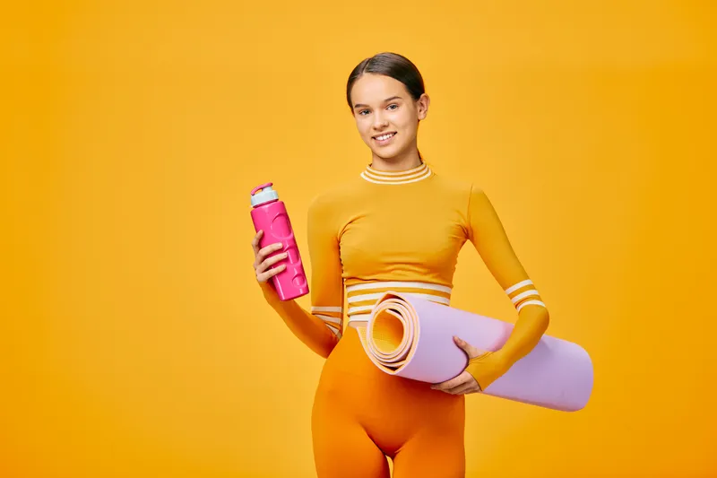 Female fitness enthusiast stands with a water bottle and yoga mat in a vivid orange studio, ready for a workout and healthy lifestyle training.