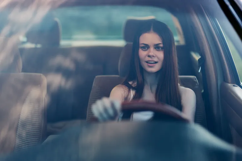 Young woman driving a car, smiling with confidence, showcasing modern fashion and vibrant emotions in a sunny setting.