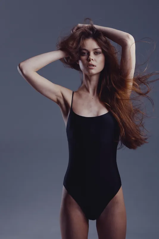 Young woman with long red hair in black bodysuit posing against dark gray background with confident expression