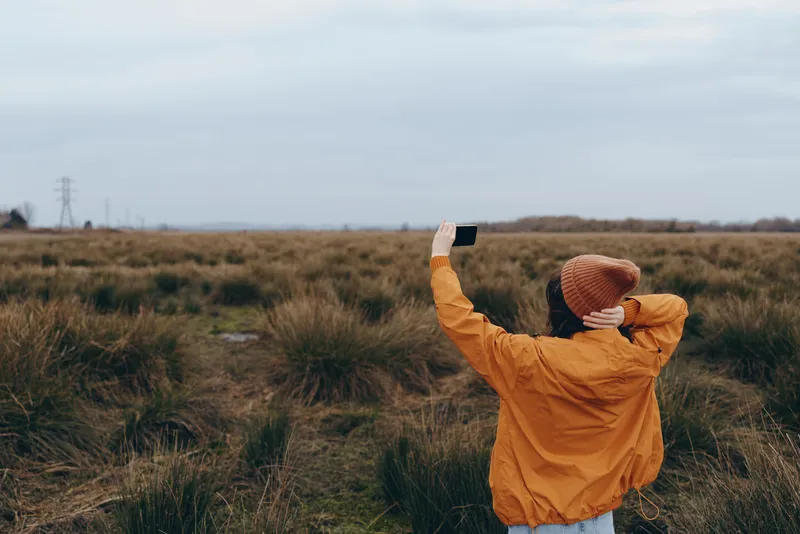 Woman lifts smartphone in autumn outdoor field, wearing coat and hat among grass, taking photo for lifestyle Natural light under cloudy sky gives serene calm mood, wide horizon background