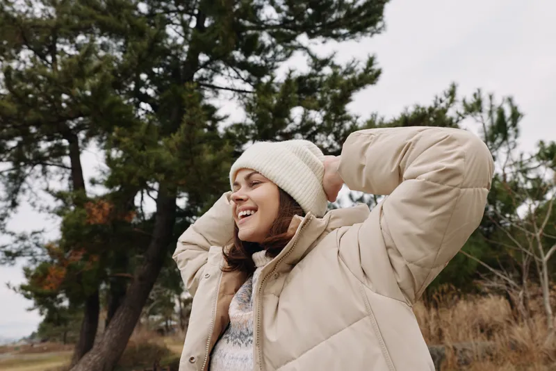 Woman in a beige puffer jacket smiles outdoors, wearing a knit beanie, posing among trees on a cool day, natural light, relaxed lifestyle fashion portrait in nature and fresh air
