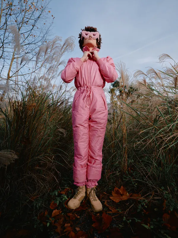Child in a pink snowsuit and owl mask stands in tall grass outdoors, posing playfully with hands near face, autumn light and clear sky creating a whimsical, creative fashion portrait in nature