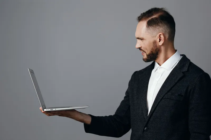 A professional man in a tailored suit holds a slim laptop against an isolated colored background, radiating confidence, focus, and a calm determination to succeed.