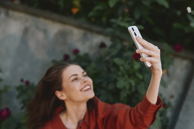 A smiling woman takes a casual selfie outdoors in daylight, capturing a sunlit moment in a garden with roses and lush greenery