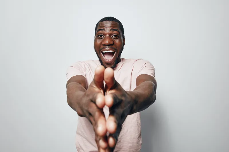 African American man with dark skin smiling and clapping hands in front of white background. Happy black male expressing excitement and joy in casual shirt.