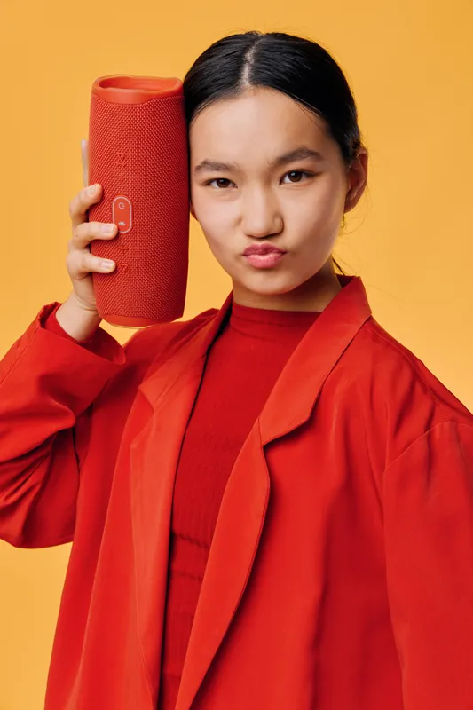 Young woman wearing red jacket holding portable wireless speaker near ear with playful expression on orange background. Technology, lifestyle and music concept with modern device and style.