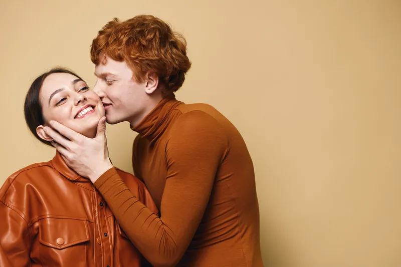 Couple in love with man kissing woman on cheek, both smiling and wearing brown tones in studio with beige background. Romantic, happy, affectionate moment captured closely.