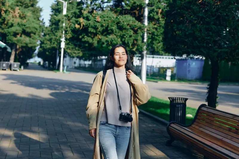 Woman strolling along a park street under daylight in a beige coat over a gray sweater and denim, camera hanging from her neck, urban casual lifestyle capture with candid mood