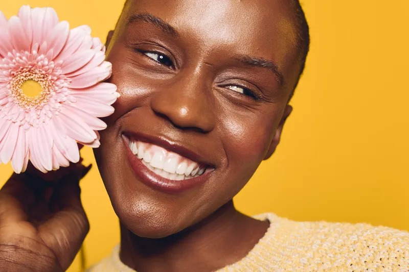 Close-up of a smiling dark skinned African woman holding a pink flower near her face against a yellow background. Concept of happiness, beauty, skincare, and natural wellness.