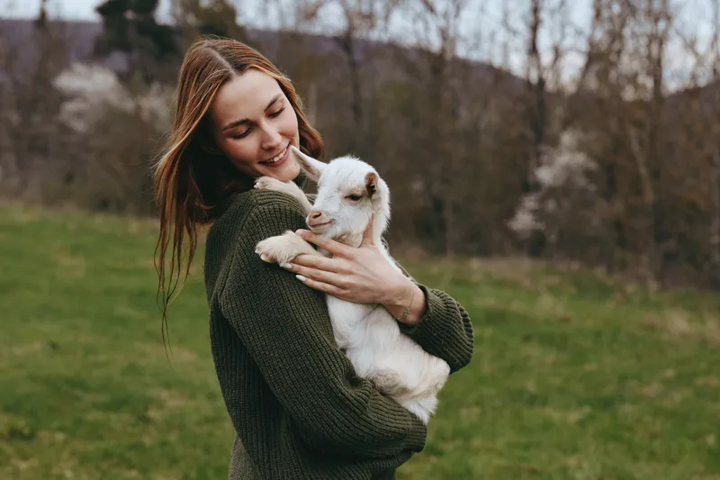 Joyful woman outdoor, gently hugging a goat kid in a green meadow, casual sweater, peaceful countryside nature, warm colors, candid lifestyle scene, connection, simple happiness and gentle breeze