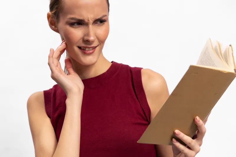 Young woman in sleeveless red top reads a book with a confused expression, touching her face. Isolated on white background. Concept for difficulty understanding, concentration, and curiosity.