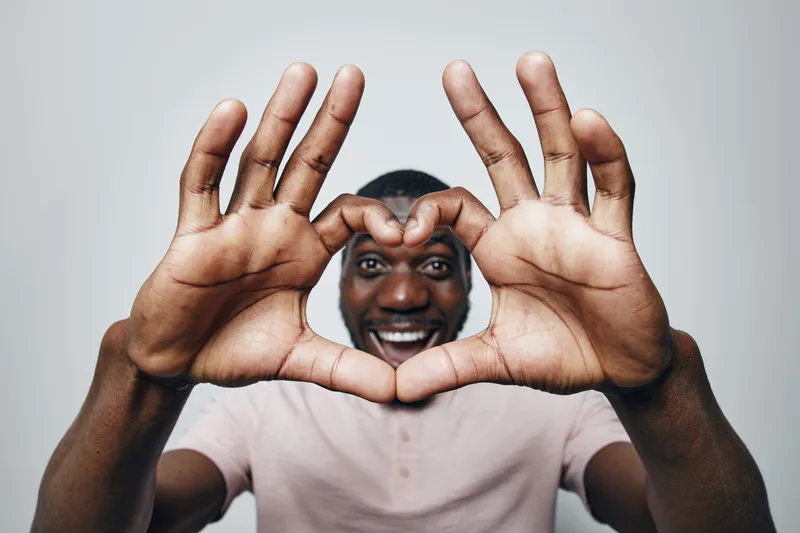 African man with dark skin making heart shape with hands close to camera, smiling and wearing light pink shirt on gray background in studio portrait.