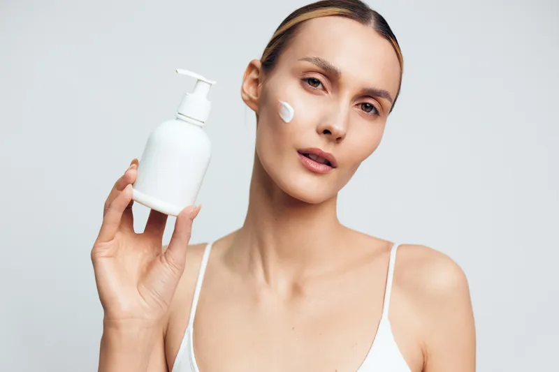 Woman models skincare routine, holding a white lotion bottle with pump, face framed by soft light, serum like moisturizer concept, beauty and hygiene, spa self care, clean minimal studio portrait