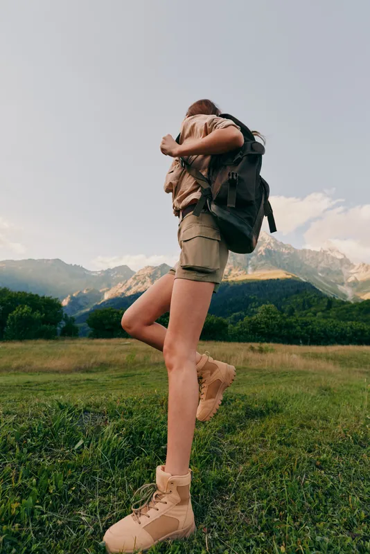 Young woman hiking outdoors in nature, wearing backpack and boots, walking on green grass with mountains in background, active lifestyle, summer adventure.