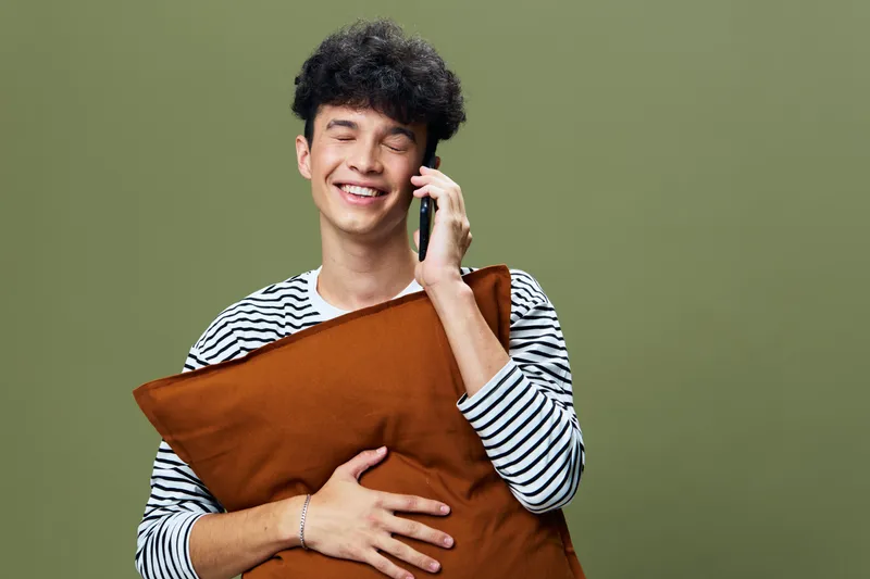 smiling young man talking on smartphone while holding brown pillow with closed eyes against green background, casual style and joyful expression