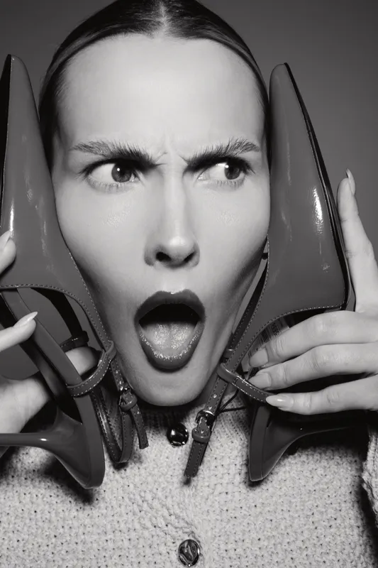 Young caucasian woman with fair skin holding high heel shoes by face, showing surprised expression in black and white.