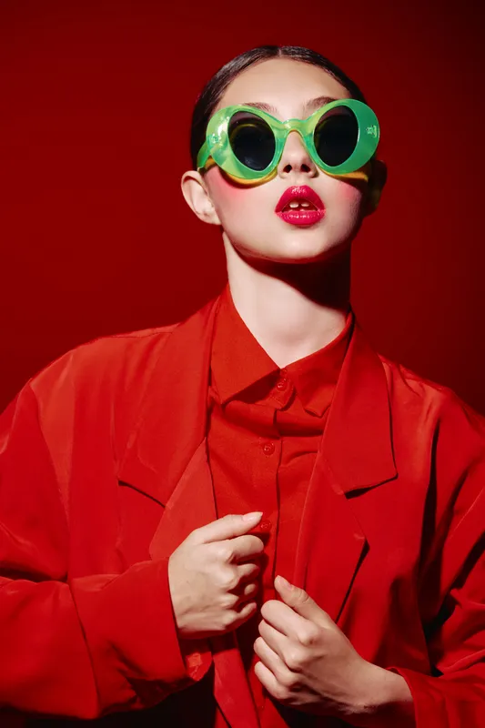 Young woman in bright red suit and bold green sunglasses poses confidently on red background in studio. Concept for beauty creativity and fashion editorial theme.