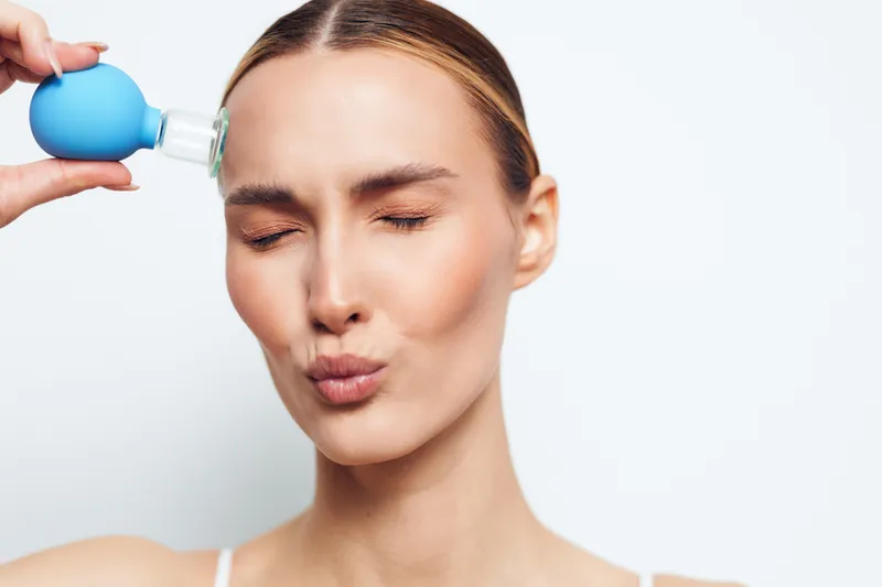 Young woman uses a facial workout suction tool pressed to her temple, eyes closed, lips puckered, demonstrating a focused beauty routine against a white background.