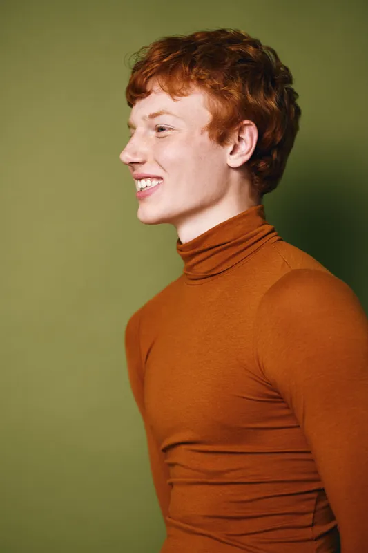 Young man with red hair wearing brown turtleneck smiling and looking sideways on green background. Portrait, casual fashion and hairstyle concept.