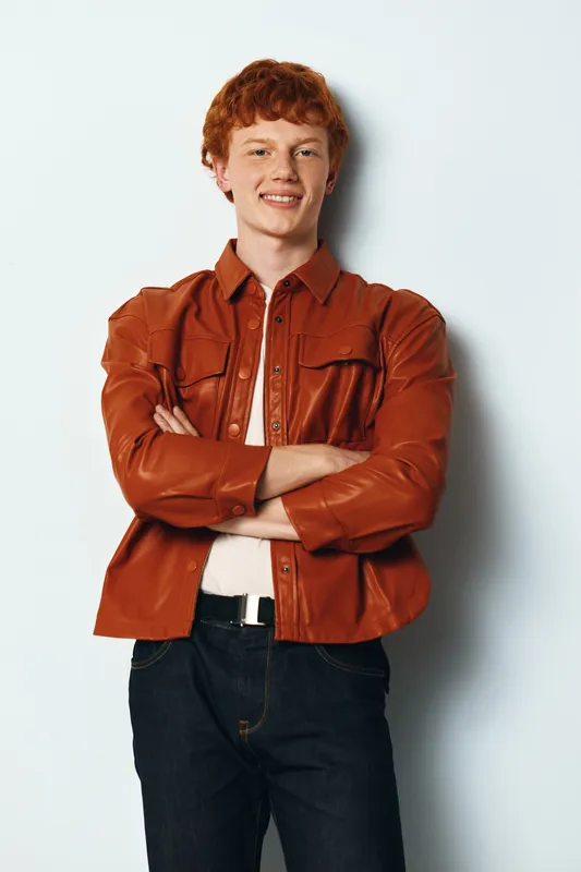 Young man with red hair standing with arms crossed smiling against white background wearing brown leather jacket and casual jeans. Portrait of confident male model.
