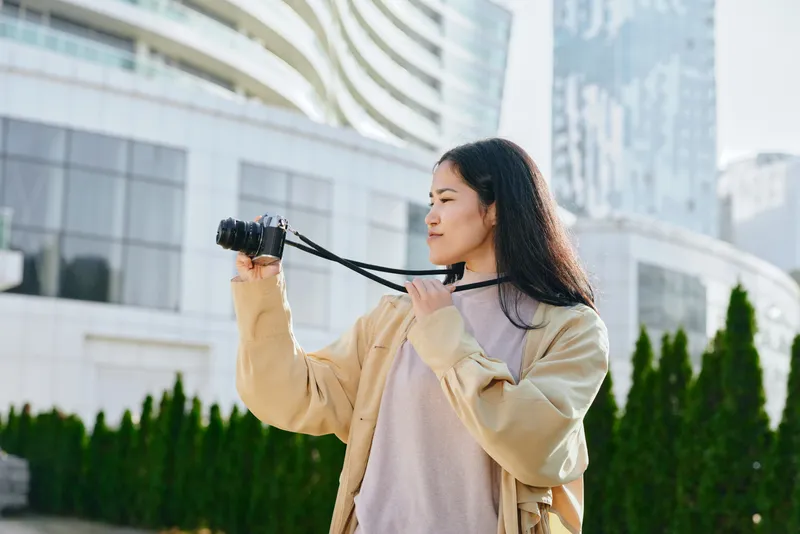 Woman with camera in hand capturing urban scenery in an outdoor setting with modern buildings, showcasing street photography style and creative observation.