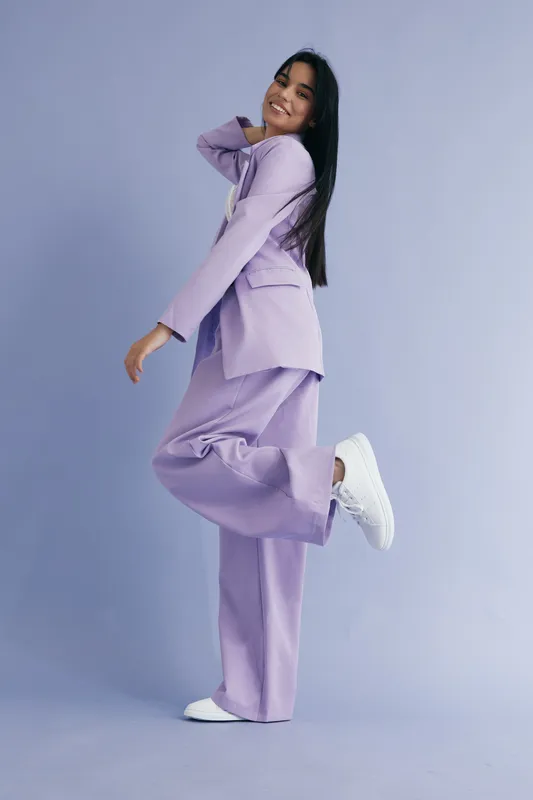 Smiling woman in violet suit posing joyfully against pastel purple background, wearing white sneakers, with long dark hair and cheerful expression, fashion and lifestyle concept.