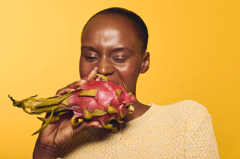African woman with dark skin bites into fresh dragon fruit, wearing a beige sweater. The bright yellow background enhances the vibrant colors, concept of healthy eating and natural fruit.