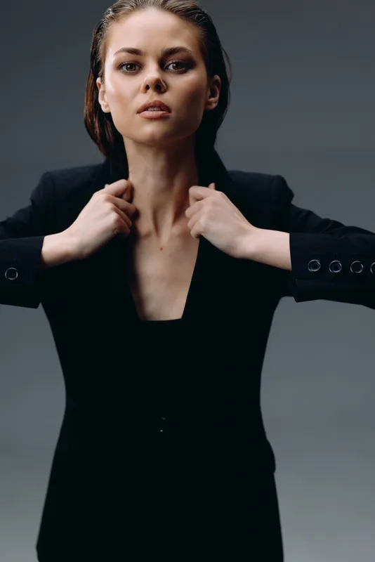 Confident woman in black suit adjusting collar on gray background with serious expression and modern style