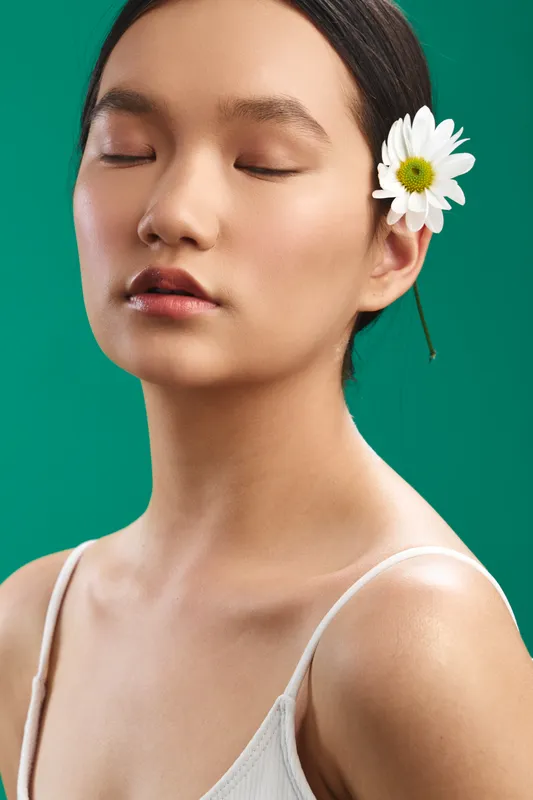 Asian woman with a tranquil expression rests with eyes closed, a flower tucked behind her ear, against a teal backdrop, conveying calm, beauty, and natural grace in a minimalist studio portrait.