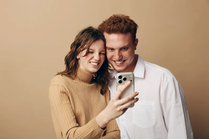 Young couple with red hair and brown hair smiling and taking selfie with smartphone on beige background. Casual clothing and happy mood create concept of friendship and modern lifestyle.