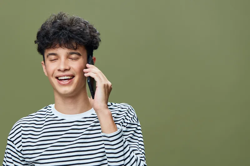 young man talking phone smiling eyes closed in striped shirt against green background, casual hairstyle, joyful expression, modern communication concept