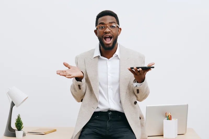 Confused excited young African American man in business casual clothing holding smartphone, gesturing with hands, puzzled facial expression, standing in modern office environment with white wall backg