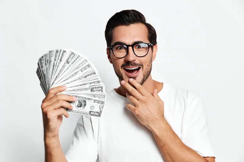 Happy cheerful man holding a fan of dollar bills while looking at the camera, showing excitement and surprise on his face against a plain white background.