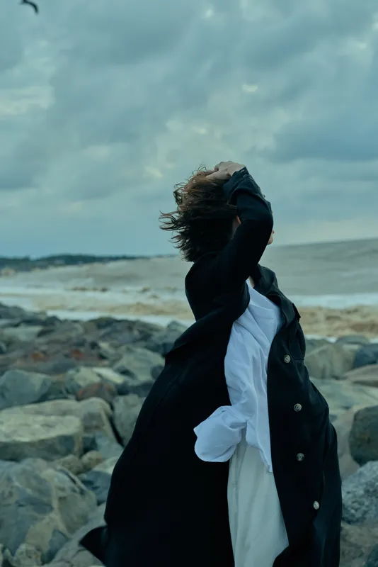 Windy coastal scene with a woman on a rocky shoreline, hair blown by gusts, wearing a dark coat and white shirt, capturing movement against a moody sea and sky