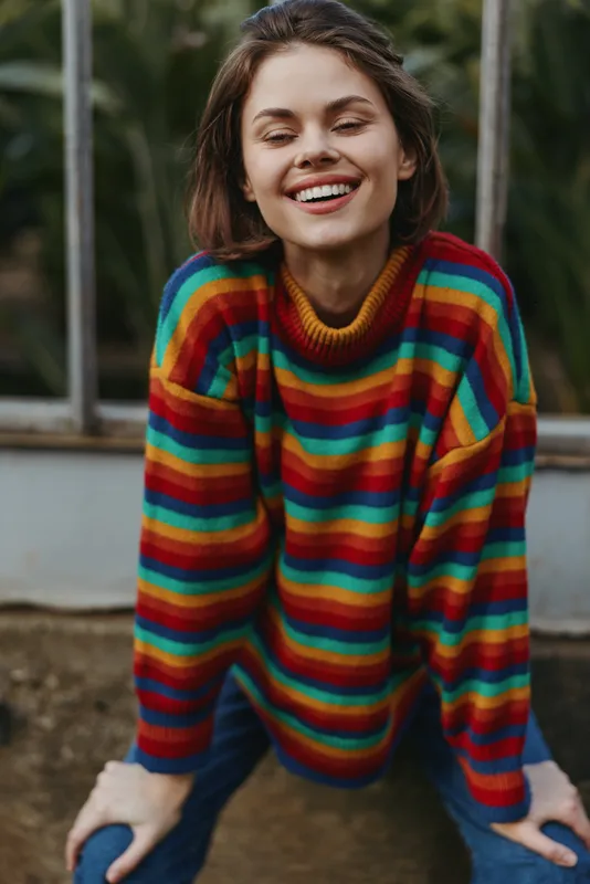 Young adult woman smiling and leaning forward in a colorful striped sweater and casual outfit with jeans in a greenhouse outdoor setting, joyful lifestyle portrait for fashion and travel.