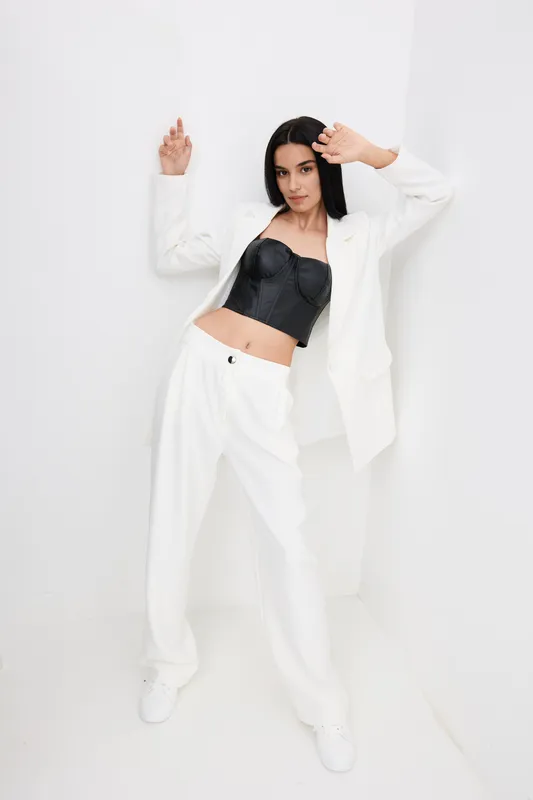 Stylish woman wearing white suit and black corset, posing confidently against a white wall in a minimalistic setting with modern fashion elements and clean lines.