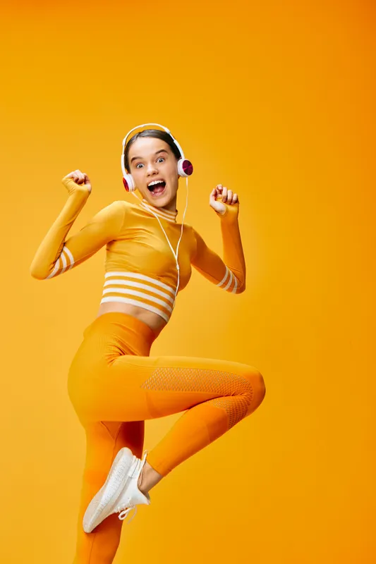 Energetic woman in bright orange workout outfit jumping mid air with headphones in a vivid orange studio, capturing dynamic fitness action, momentum, and style for sports and lifestyle marketing.