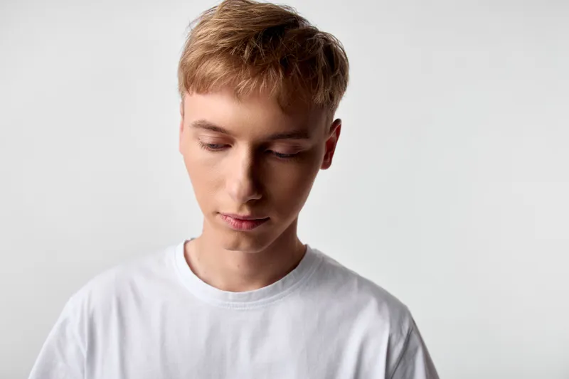 Young man contemplation downward gaze, white shirt, calm mood, minimal studio, quiet focus, thoughtful expression, soft lighting, introspection for lifestyle, fashion, or editorial use
