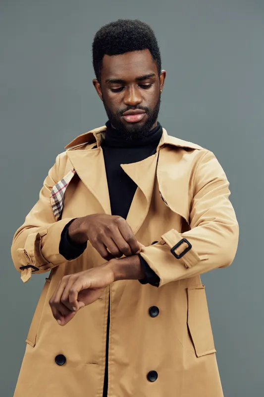 Stylish young African man adjusting his coat sleeve, exuding confidence and elegance against a muted green background Fashion, personality, confidence, and elegance stand out