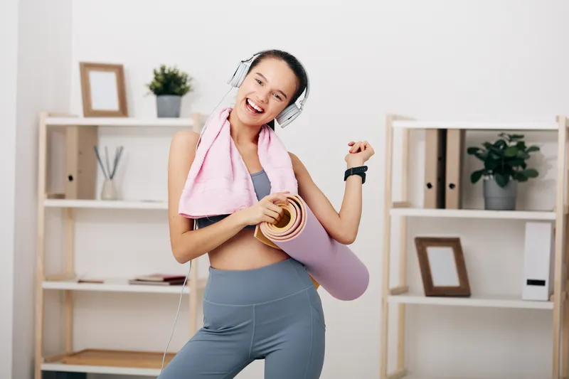 Fitness oriented scene with a cheerful woman at home, holding a yoga mat and roll, wearing headphones and a towel draped over neck, showcasing energetic workout and healthy lifestyle.