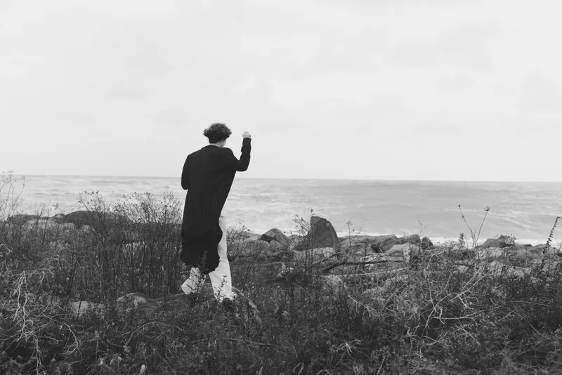 Black and white image of a lone walker along a rocky coast, waving toward the horizon, with a calm sea and windswept sparse shoreline
