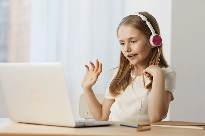 Young girl participating in an online class with headphones, cheerful and engaged, in a bright indoor setting, showcasing modern learning and technology