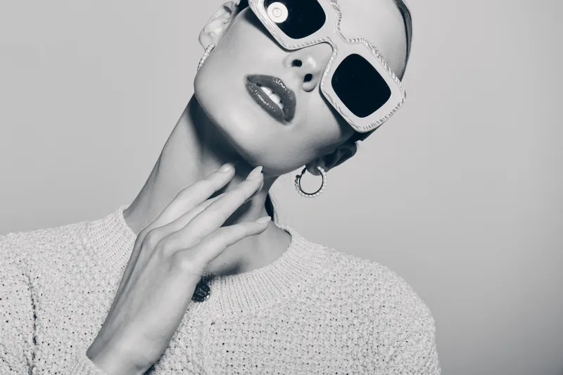 Stylish white woman with fair skin wears large sunglasses and textured sweater. Close-up portrait with glossy lips and hoop earrings in black and white. Concept of fashion and elegance.