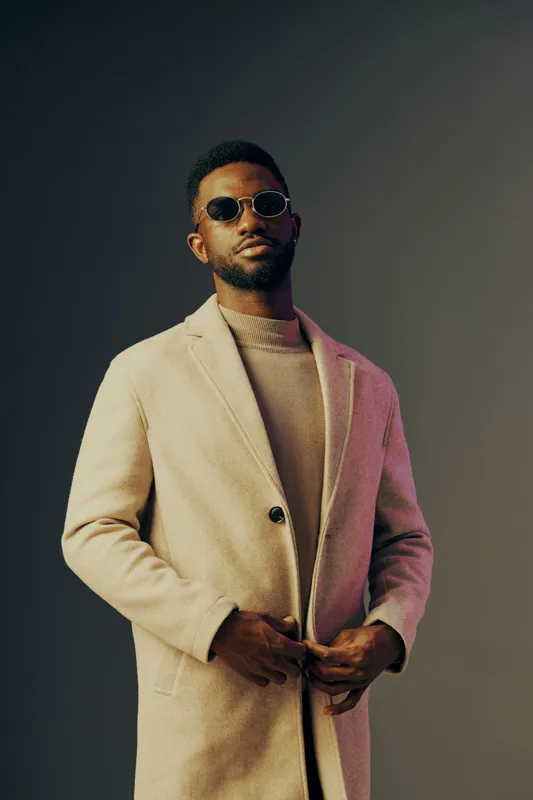 Stylish Black man in a beige coat and sunglasses against a minimalist gray background, exuding confidence and modern fashion trends